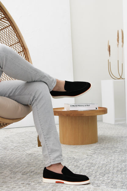 Heritage Suède Loafer Zwart – gecombineerd met een zakelijke outfit op een stenen achtergrond.